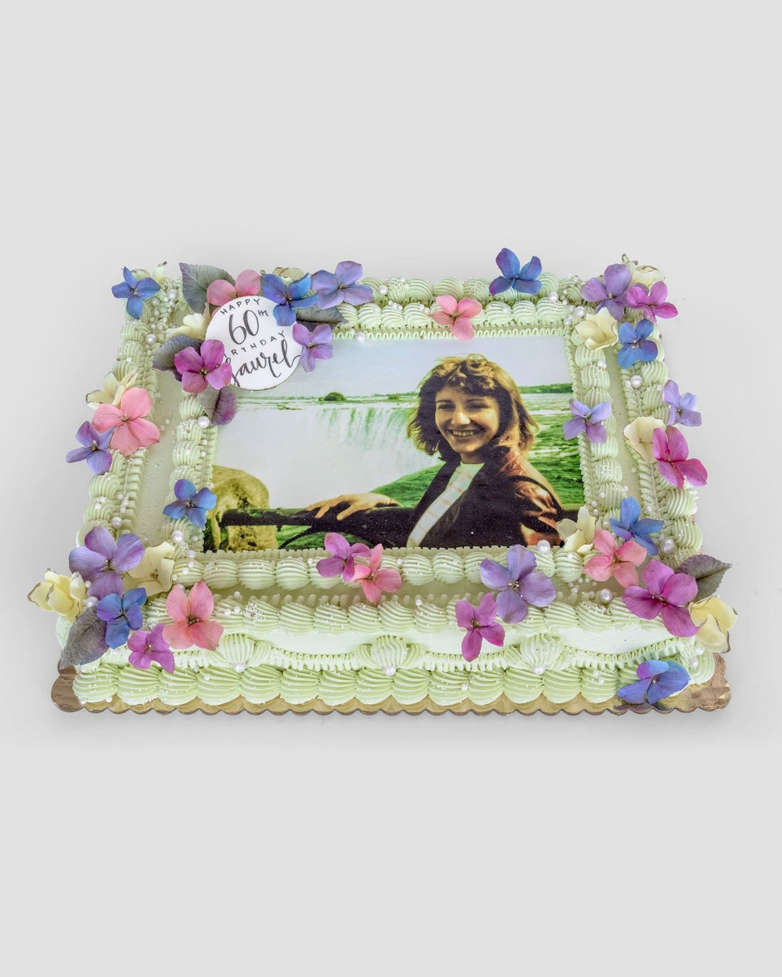 The Sugar Flower Sheet Cake by Jenna Rae Cakes is a rectangular cake adorned with pink, purple, and blue sugar flowers, piped cream borders, and an edible outdoor photo of a smiling woman with a 60 & proud topper.