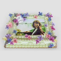 The Sugar Flower Sheet Cake by Jenna Rae Cakes is a rectangular cake adorned with pink, purple, and blue sugar flowers, piped cream borders, and an edible outdoor photo of a smiling woman with a 60 & proud topper.