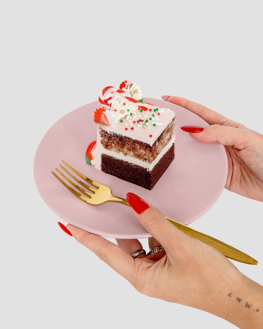 A person with red nails holds a pink plate with a slice of Jenna Rae Cakes’ Sugar Flower Sheet Cake, topped with white frosting, sprinkles, and candy, while holding a gold fork in the other hand.