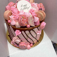 The Full Works Cake by Jenna Rae Cakes: a pink and white birthday cake topped with pink macarons, meringues, candies, sprinkles, chocolate drip, a happy birthday! topper, and set on a gold base.