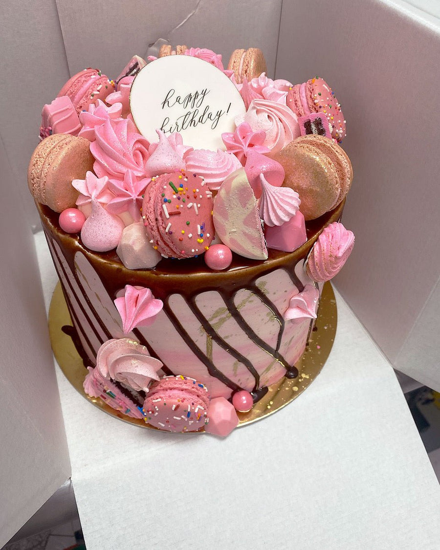 The Full Works Cake by Jenna Rae Cakes: a pink and white birthday cake topped with pink macarons, meringues, candies, sprinkles, chocolate drip, a happy birthday! topper, and set on a gold base.