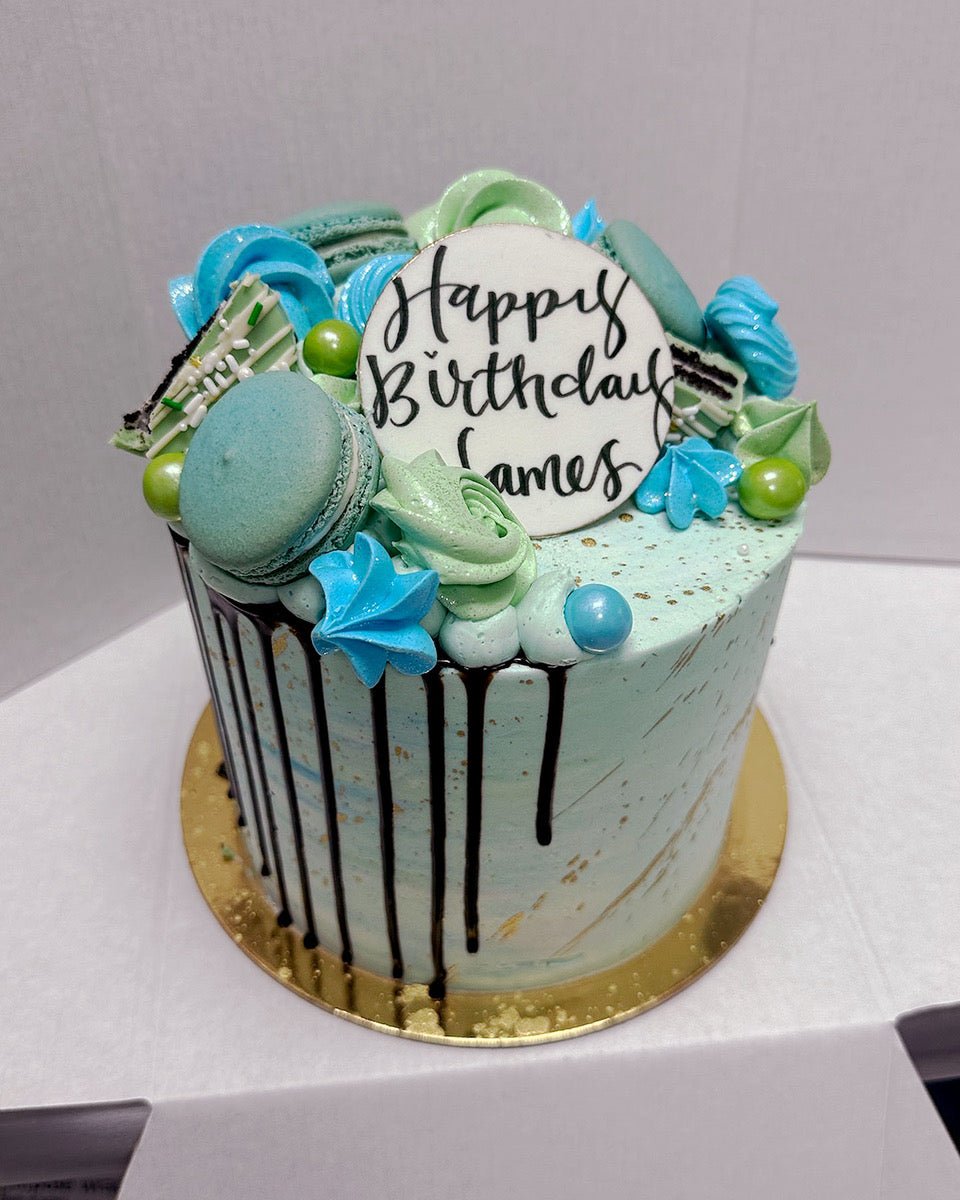 The Half Works Cake by Jenna Rae Cakes is a blue and green birthday cake with macarons, meringues, chocolate drizzle, and a white Happy Birthday James disc, presented on a gold board in a white box.