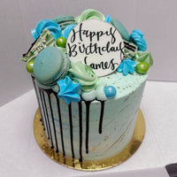 The Half Works Cake by Jenna Rae Cakes is a blue and green birthday cake with macarons, meringues, chocolate drizzle, and a white Happy Birthday James disc, presented on a gold board in a white box.