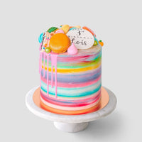 The Half Works Cake by Jenna Rae Cakes features a tall, colorful watercolor design with pink drip icing, topped with assorted macarons, meringues, and a white disc reading 3rd birthday Boris, displayed on a marble cake stand.