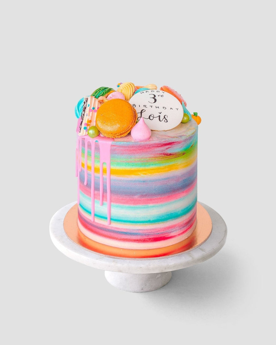 The Half Works Cake by Jenna Rae Cakes features a tall, colorful watercolor design with pink drip icing, topped with assorted macarons, meringues, and a white disc reading 3rd birthday Boris, displayed on a marble cake stand.