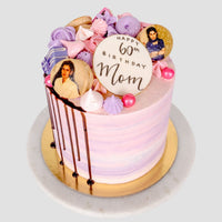 The Half Works Cake by Jenna Rae Cakes features pink and purple layers, meringues, photo cookies, pink pearls, a “Happy 60th Birthday Mom” cookie, and chocolate drip on the side, displayed on a white and gold cake stand.