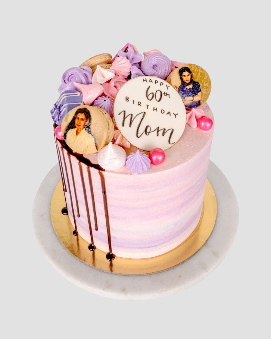 The Half Works Cake by Jenna Rae Cakes features pink and purple layers, meringues, photo cookies, pink pearls, a “Happy 60th Birthday Mom” cookie, and chocolate drip on the side, displayed on a white and gold cake stand.