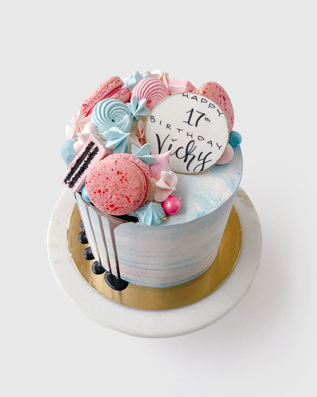 The Half Works Cake by Jenna Rae Cakes is a pastel birthday cake topped with pink and blue macarons, meringues, chocolate drizzle, and a white “Happy 17th Birthday Vicky” topper, served on a marble stand.