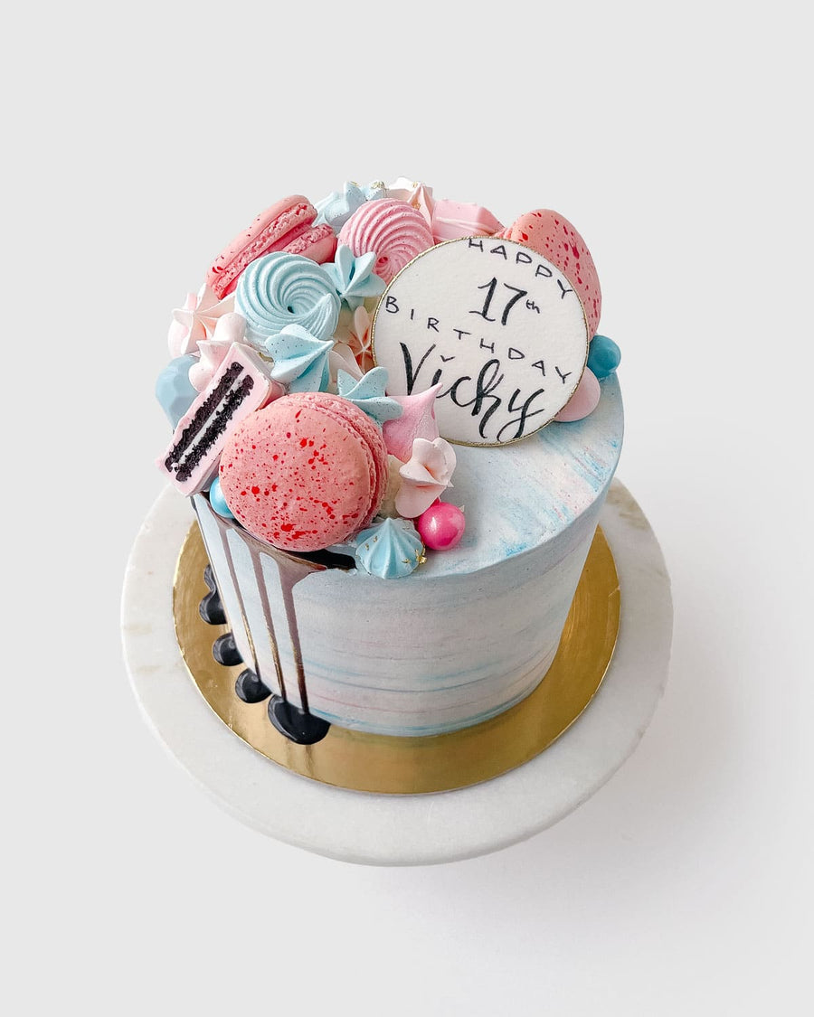 The Half Works Cake by Jenna Rae Cakes is a pastel birthday cake topped with pink and blue macarons, meringues, chocolate drizzle, and a white “Happy 17th Birthday Vicky” topper, served on a marble stand.