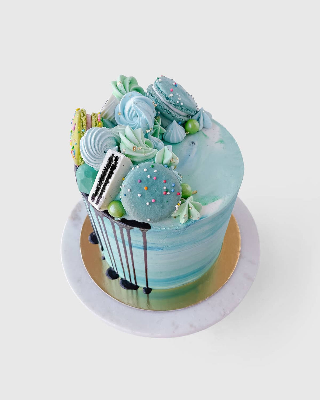 The Half Works Cake by Jenna Rae Cakes features a blue and white marbled design topped with blue and green macarons, meringues, and colorful sprinkles on a gold and marble cake board.