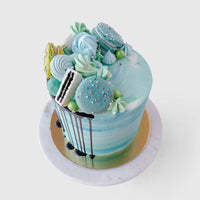 The Half Works Cake by Jenna Rae Cakes features a blue and white marbled design topped with blue and green macarons, meringues, and colorful sprinkles on a gold and marble cake board.
