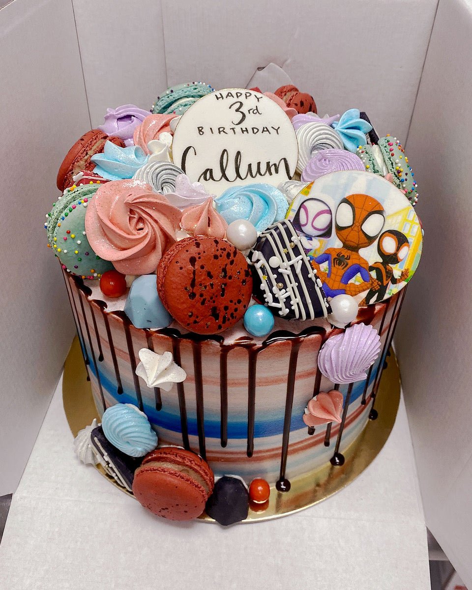 The Kids Works Cake by Jenna Rae Cakes is a colorful birthday cake topped with meringues, macarons, candies, chocolate drip, and a Spider-Man cookie. “Happy 3rd Birthday Callum” is written in black icing on top.
