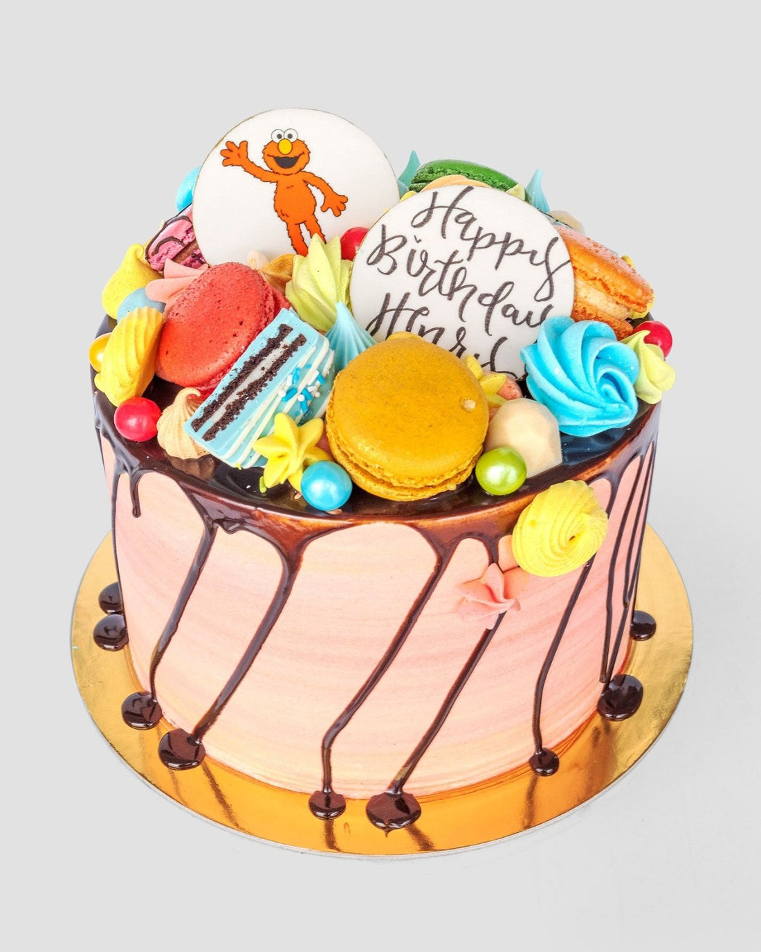 The Kids Works Cake by Jenna Rae Cakes features pink frosting, chocolate drizzle, macarons, meringues, candies, and two cookie toppers—a cartoon character and a Happy Birthday design—making it a colorful birthday cake.