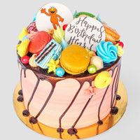 The Kids Works Cake by Jenna Rae Cakes features pink frosting, chocolate drizzle, macarons, meringues, candies, and two cookie toppers—a cartoon character and a Happy Birthday design—making it a colorful birthday cake.