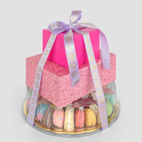 The Treat Tower by Jenna Rae Cakes features two pink gift boxes with a purple ribbon atop a clear round container filled with colorful macarons, all beautifully arranged on a gold tray against a plain light background.