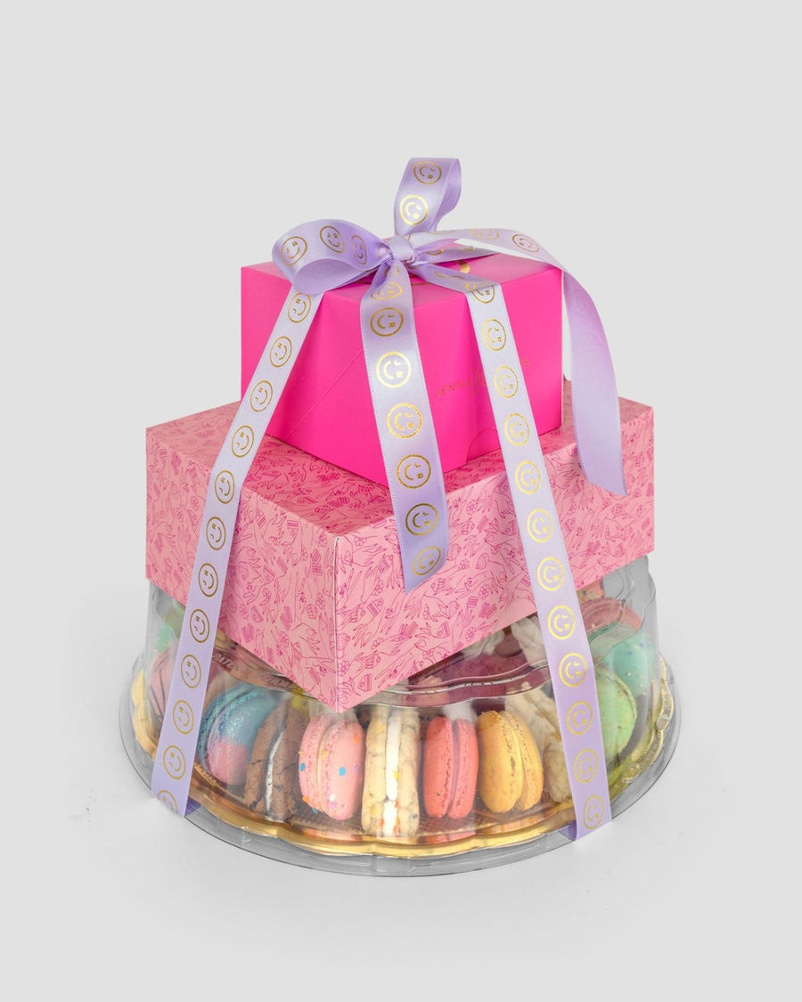 The Treat Tower by Jenna Rae Cakes features two pink gift boxes with a purple ribbon atop a clear round container filled with colorful macarons, all beautifully arranged on a gold tray against a plain light background.