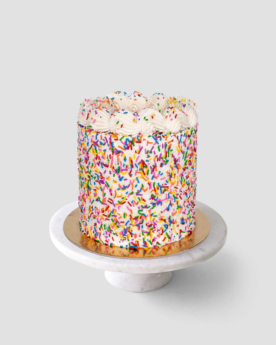 The Vanilla Sprinkle Cake by Jenna Rae Cakes is a round cake covered in colorful rainbow sprinkles with white frosting swirls, displayed on a white marble cake stand against a plain light gray background.