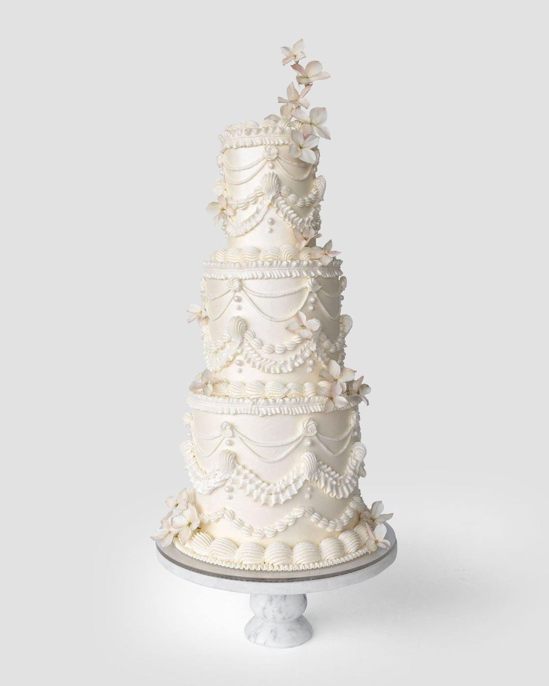 The Wedding Cakes $1,000+ by Jenna Rae Cakes features four tiers of white cake with piped icing, scalloped designs, and delicate sugar flowers, showcased on a marble stand against a simple light backdrop.