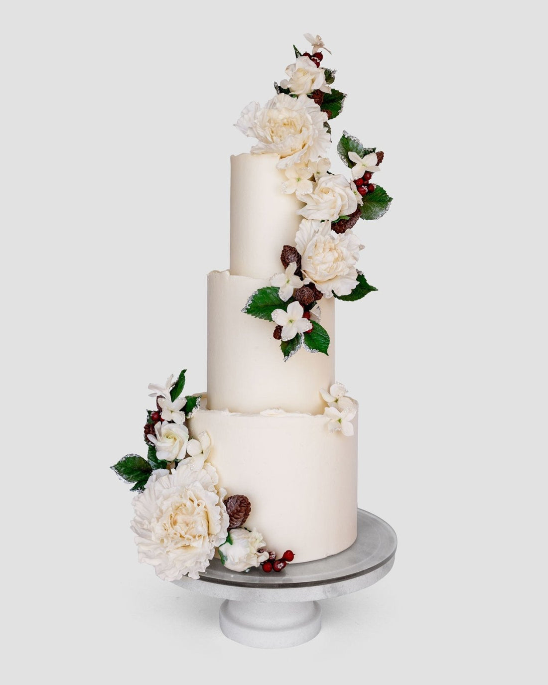 A three-tiered white cake decorated with white flowers, green leaves, and small red berries, displayed on a white cake stand against a plain background.