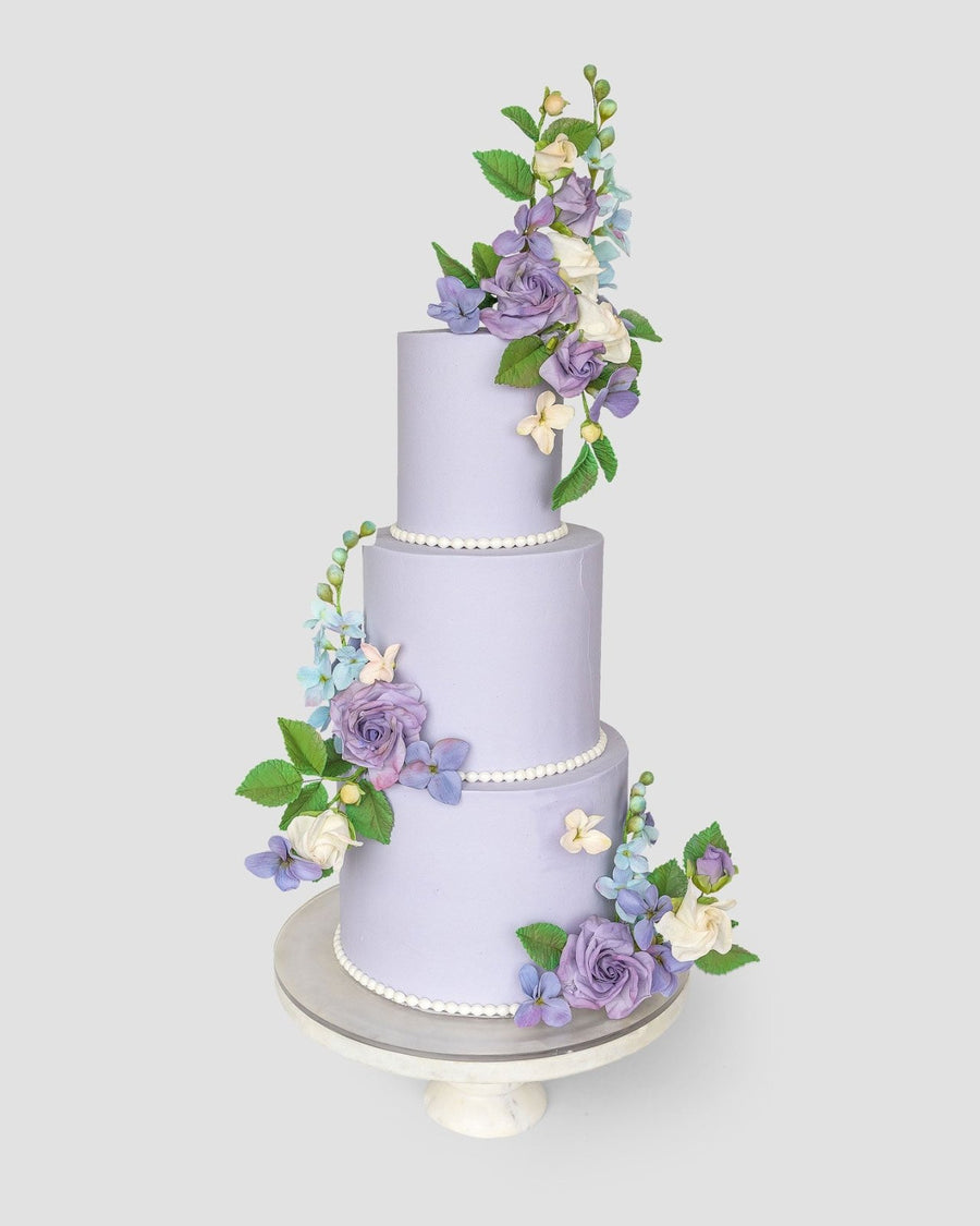 A three-tiered lavender Wedding Cake by Jenna Rae Cakes ($1,000+) features lifelike purple, blue, and white sugar flowers with green leaves, presented on a white cake stand against a simple light backdrop.