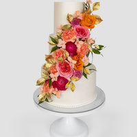 Three-tiered white cake decorated with vibrant pink, orange, and peach flowers, green leaves, and gold-painted leaves cascading down the side. The cake sits on a white pedestal stand against a light background.