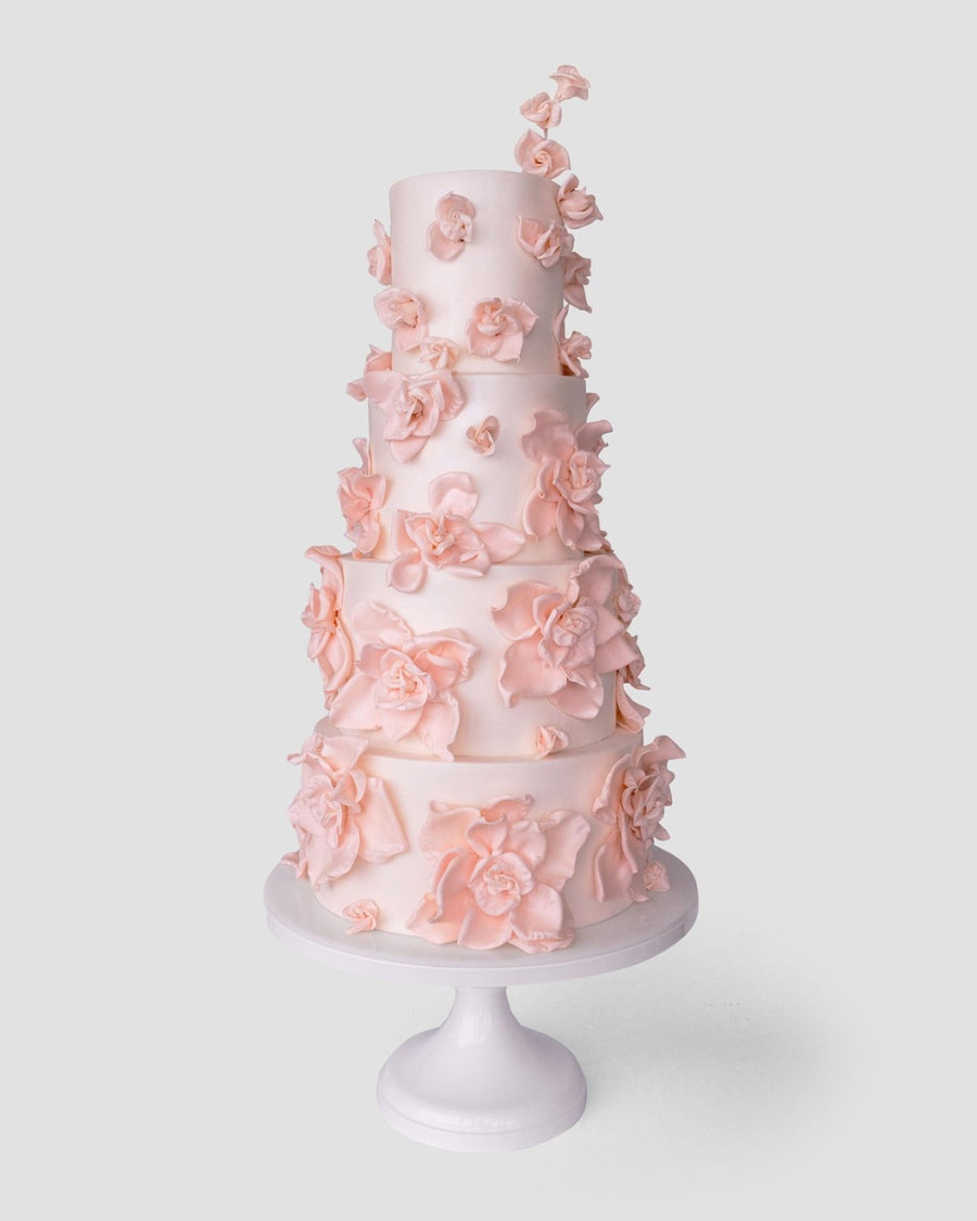 A tall, five-tier white wedding cake by Jenna Rae Cakes ($1,000+) features delicate light pink sugar flowers and is displayed on a white stand against a plain light background.