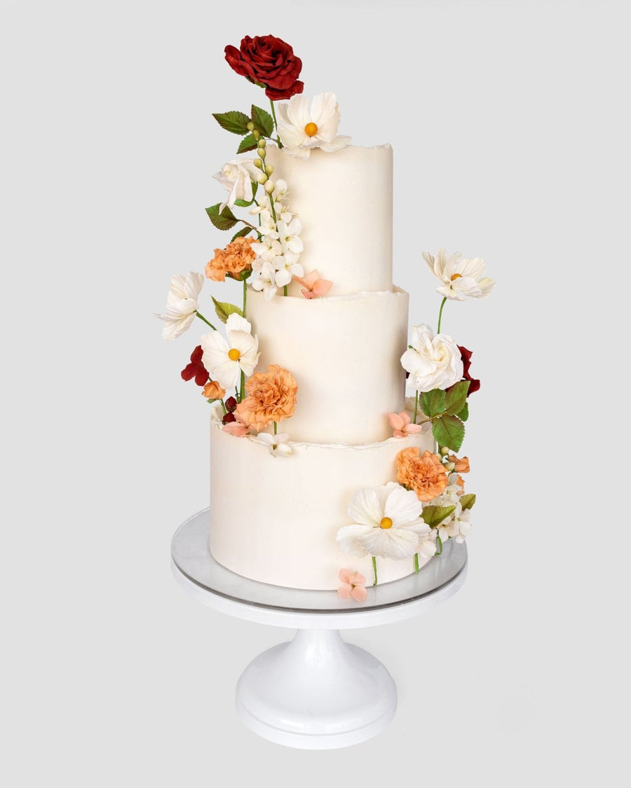 A three-tiered white Wedding Cakes $1,000+ by Jenna Rae Cakes, adorned with realistic red, orange, and white sugar flowers, is displayed on a white cake stand against a plain background.