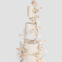 A four-tiered white wedding cake with floating tiers, decorated with cascading delicate white and blush pink sugar flowers and leaves, set against a plain light gray background.