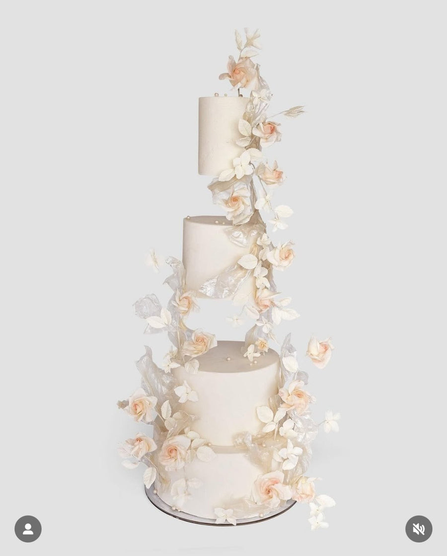 A four-tiered white wedding cake with floating tiers, decorated with cascading delicate white and blush pink sugar flowers and leaves, set against a plain light gray background.