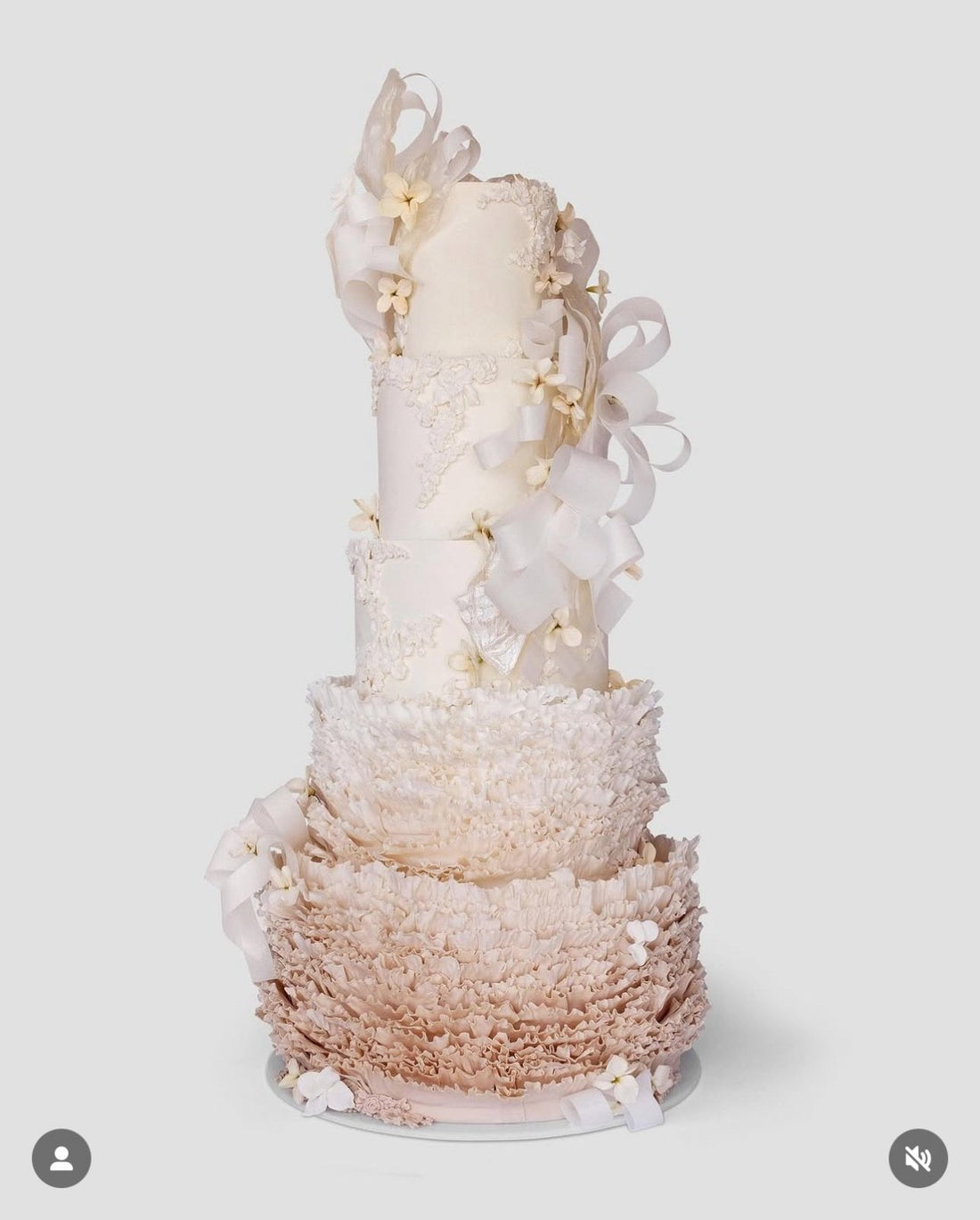 A tall, elegant tiered cake decorated with delicate white bows, small white flowers, and intricate ruffled icing in soft shades of white and beige, set against a plain light background.
