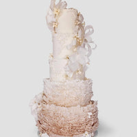 A tall, elegant tiered cake decorated with delicate white bows, small white flowers, and intricate ruffled icing in soft shades of white and beige, set against a plain light background.