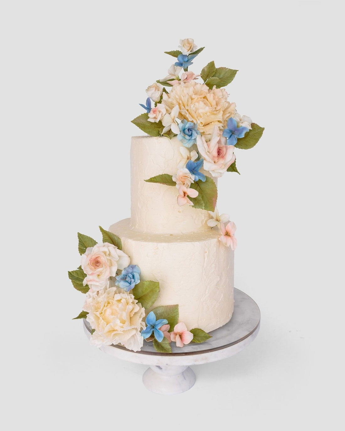 Jenna Rae Cakes Wedding Cakes Under $1,000 features a two-tiered white cake adorned with pastel pink, blue, and white flowers plus green leaves, displayed on a marble stand against a plain light background.