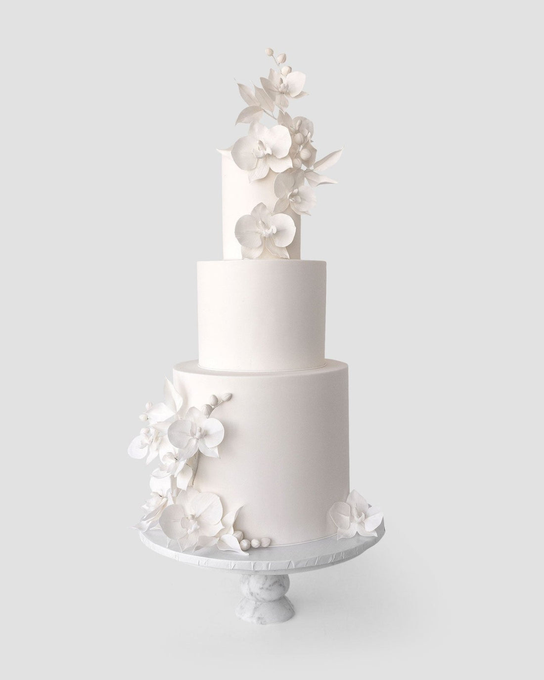 A three-tiered white cake from Jenna Rae Cakes Wedding Cakes Under $1,000 collection features elegant sugar flowers and leaves, displayed on a pedestal cake stand against a light gray background.