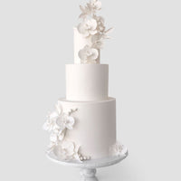 A three-tiered white cake from Jenna Rae Cakes Wedding Cakes Under $1,000 collection features elegant sugar flowers and leaves, displayed on a pedestal cake stand against a light gray background.