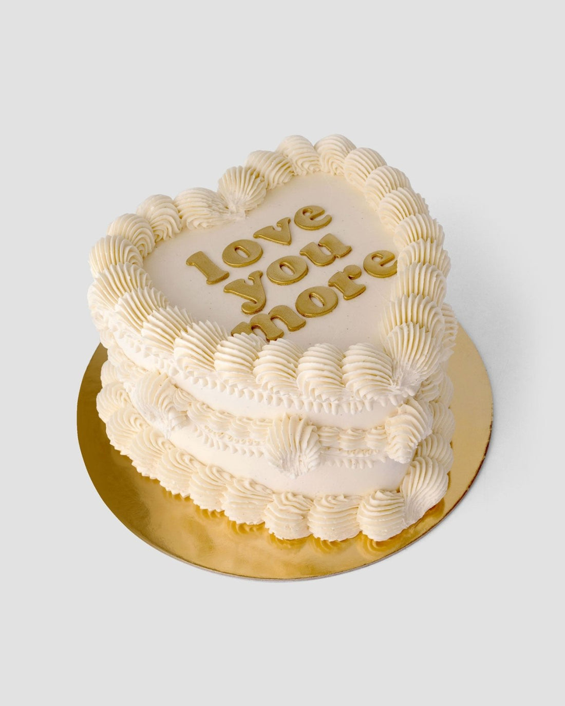 A heart-shaped cake from Jenna Rae Cakes’ Wedding Cakes Under $200 features white frosting, piped edges, and love you more in gold on top, set on a gold board against a simple backdrop.