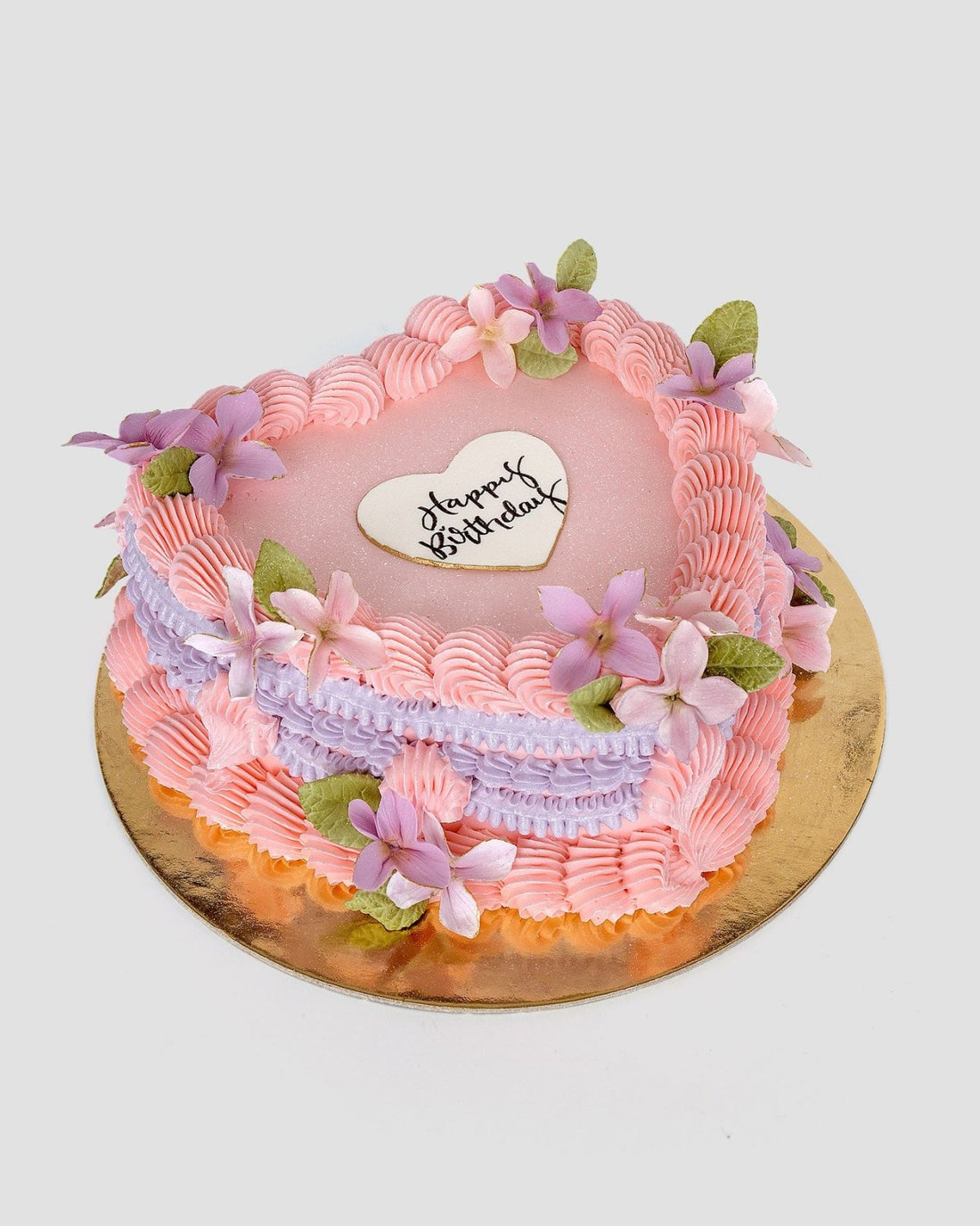 Jenna Rae Cakes’ Wedding Cakes Under $200 features a heart-shaped design with pink and purple frosting, purple flowers, green leaves, and a white “Happy Birthday” topper on a gold board.