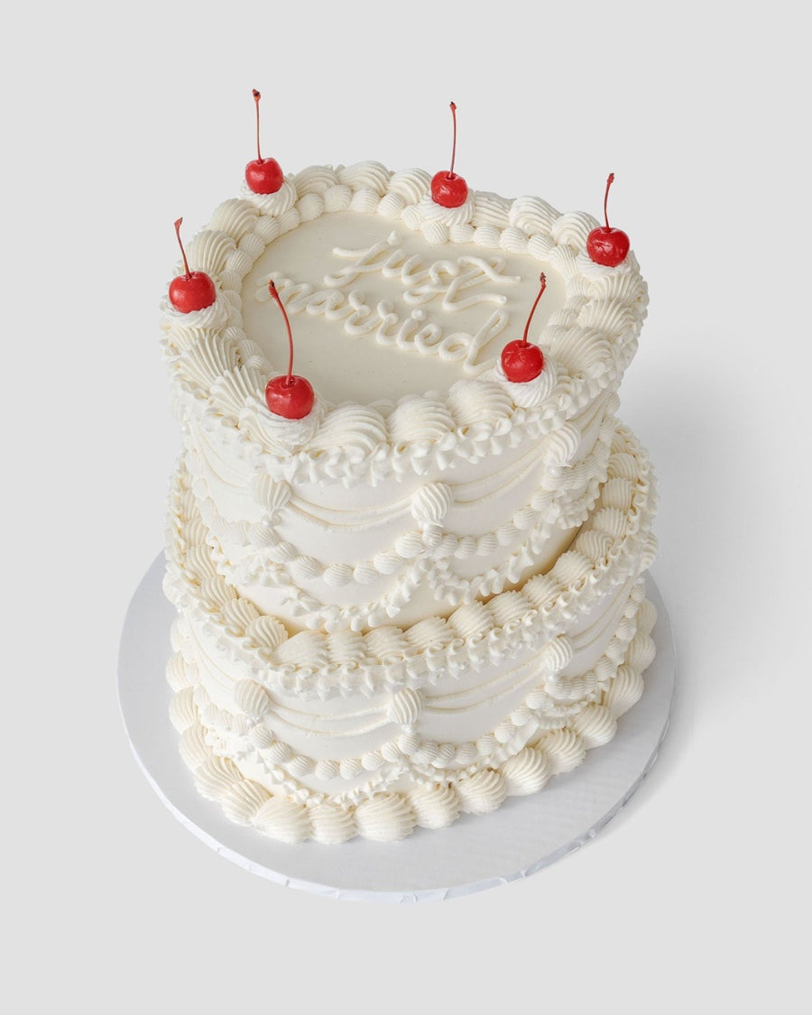 The “Wedding Cakes Under $500” by Jenna Rae Cakes is a two-tiered white frosted cake with piped cream designs, six red cherries on top, and cursive writing on the upper tier, presented on a white cake board.