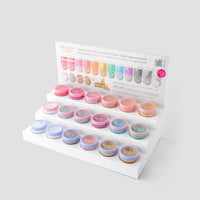 The White Acrylic Dazzle Dust Stand by Jenna Rae Cakes holds small round jars of shimmer powders, with three tiers and a sign at the back showing color options and product info. Package includes 1 stand.