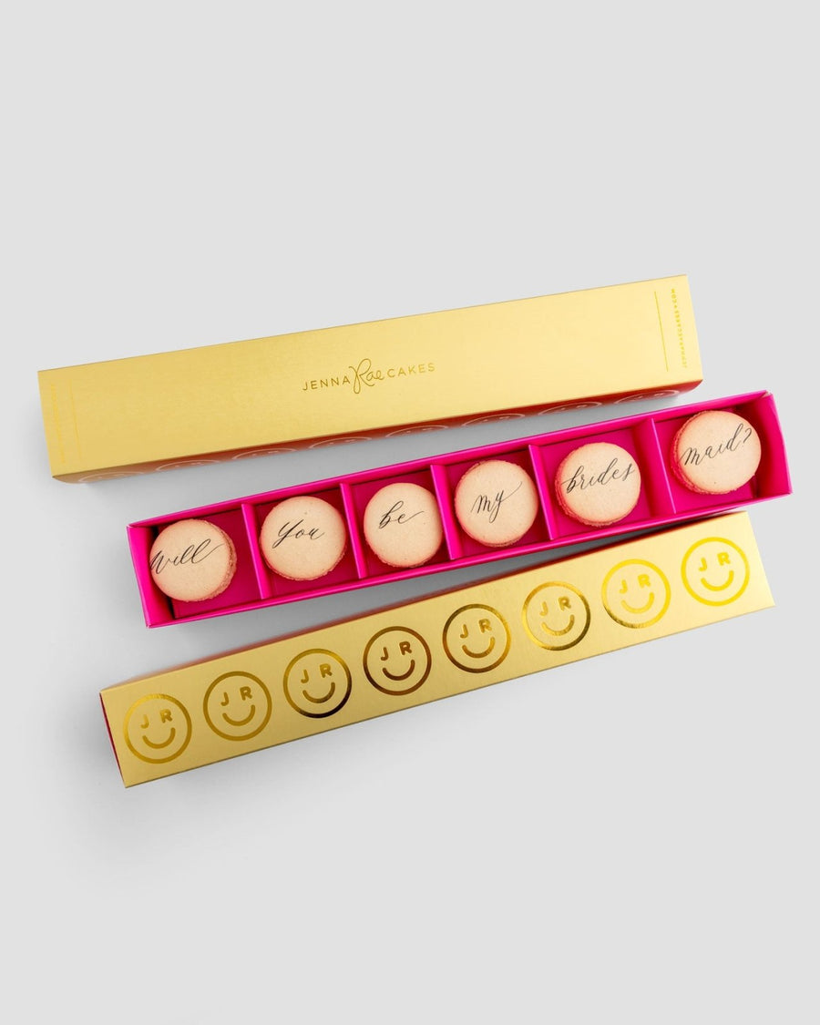 The Will You Be My... Gift Box by Jenna Rae Cakes features six macarons with “Will you be my bridesmaid?” in cursive, plus an open box with six smiley face and JR initial macarons, all set against a light gray background.