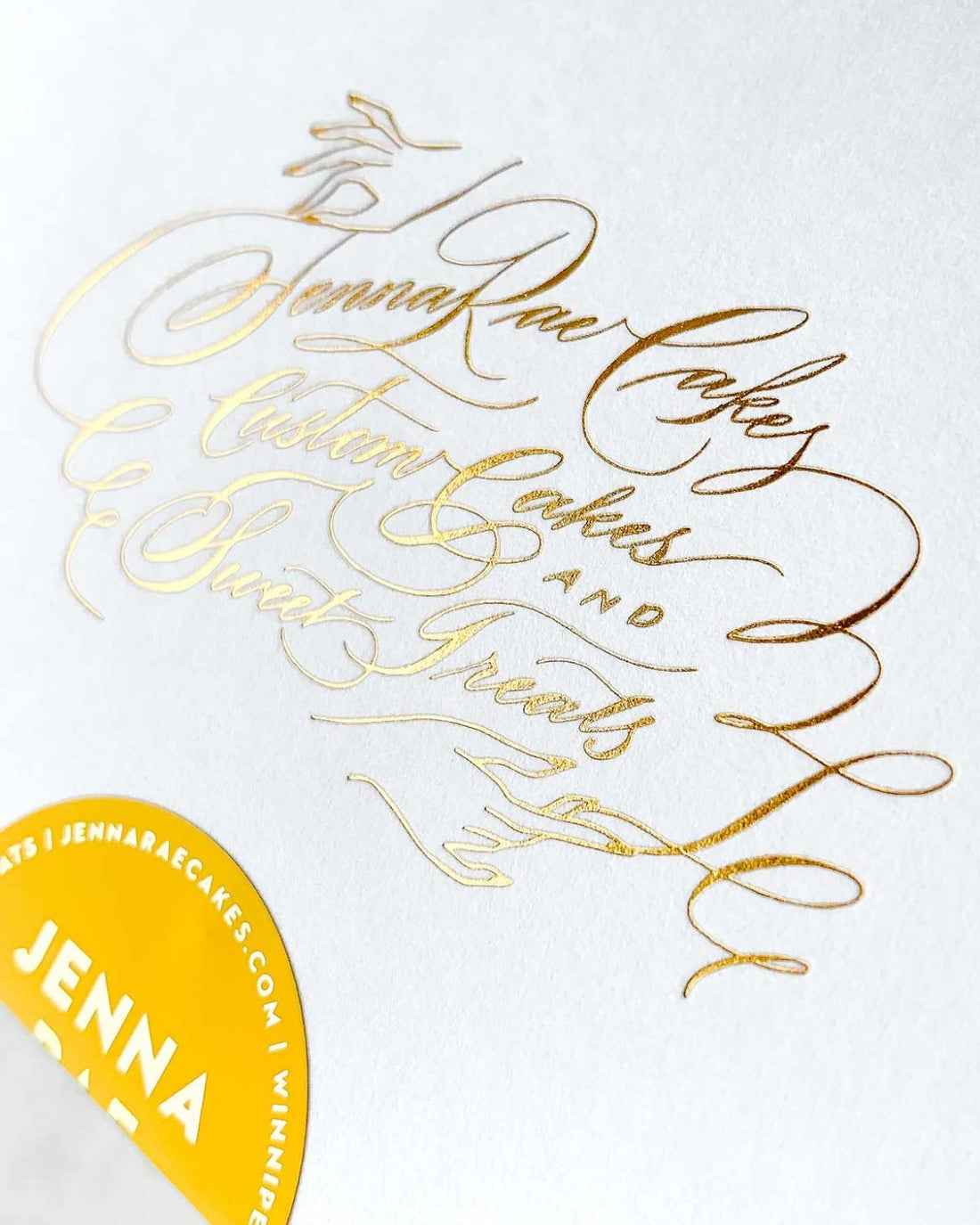 A white card with elegant gold cursive lettering reads Jenna Rae Cakes. In the bottom left, a partial yellow sticker shows the name Jenna Rae. Displayed with the Cookie Sandwich Gift Box from Jenna Rae Cakes.