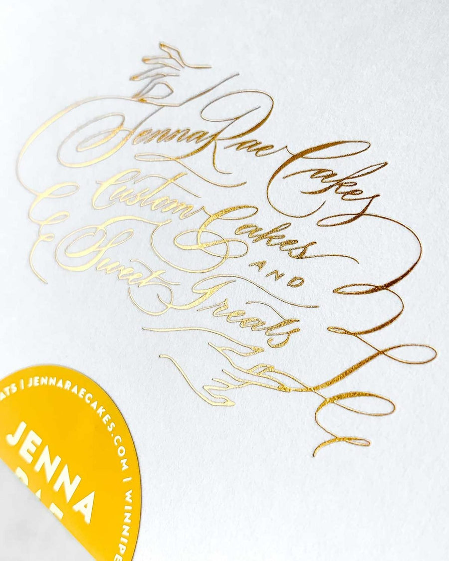 A white card with elegant gold cursive lettering reads Jenna Rae Cakes. In the bottom left, a partial yellow sticker shows the name Jenna Rae. Displayed with the Cookie Sandwich Gift Box from Jenna Rae Cakes.