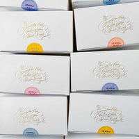 A stack of white Cookie Sandwich Gift Boxes by Jenna Rae Cakes, each featuring elegant gold script and a round sticker—pink, yellow, blue, or orange—indicating different flavors.