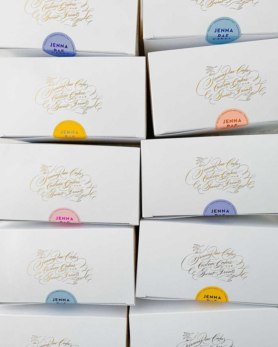 A stack of white Cookie Sandwich Gift Boxes by Jenna Rae Cakes, each featuring elegant gold script and a round sticker—pink, yellow, blue, or orange—indicating different flavors.