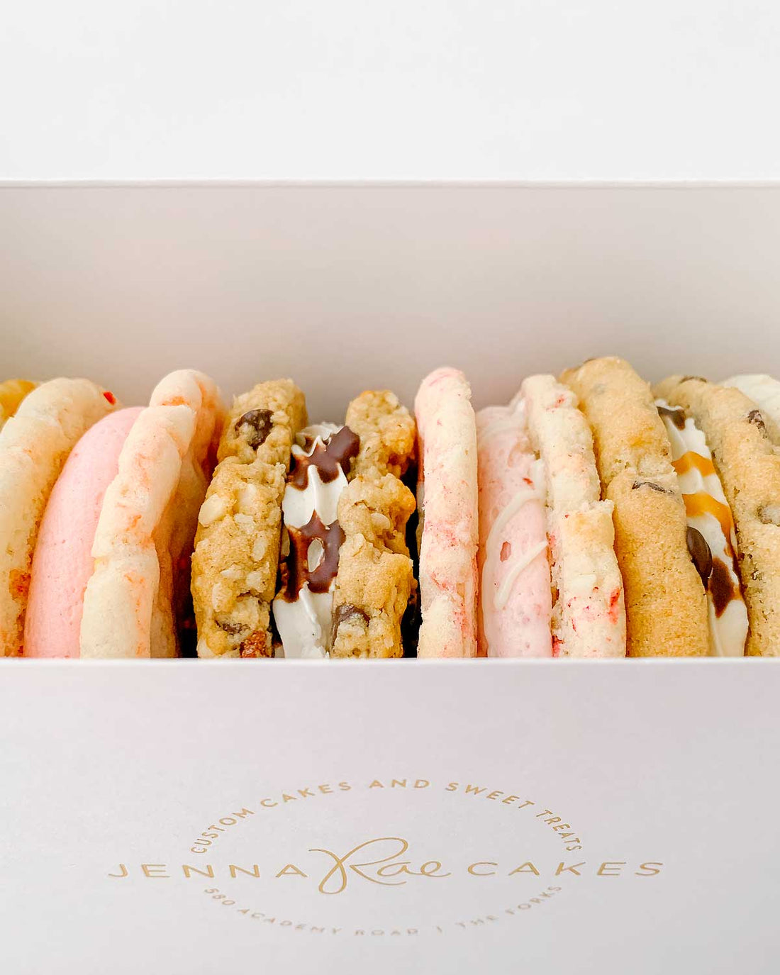 The Jenna Rae Cakes Cookie Sandwich Gift Box features a white box with the brands logo, filled with assorted cookie sandwiches in various flavors and colors, some cream-filled or drizzled with chocolate.