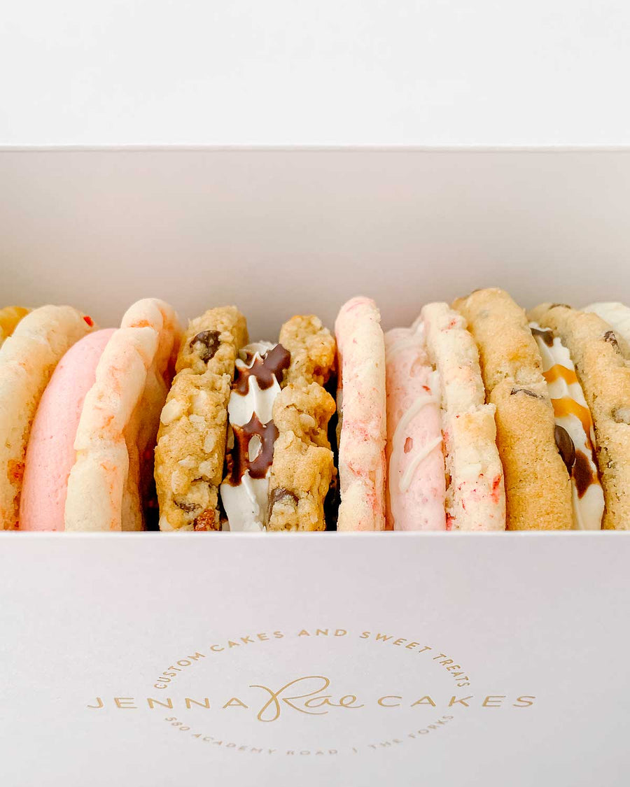 The Jenna Rae Cakes Cookie Sandwich Gift Box features a white box with the brands logo, filled with assorted cookie sandwiches in various flavors and colors, some cream-filled or drizzled with chocolate.