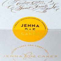 A white box labeled “Cookie Sandwich Gift Box” from Jenna Rae Cakes, featuring elegant script and a round yellow sticker with branding and website details.