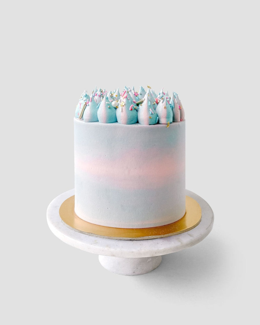 The Cotton Candy Cake by Jenna Rae Cakes is a tall, round cake with pastel blue and pink frosting, swirled dollops, and colorful sprinkles. It sits on a gold cake board and white marble stand against a plain gray background.