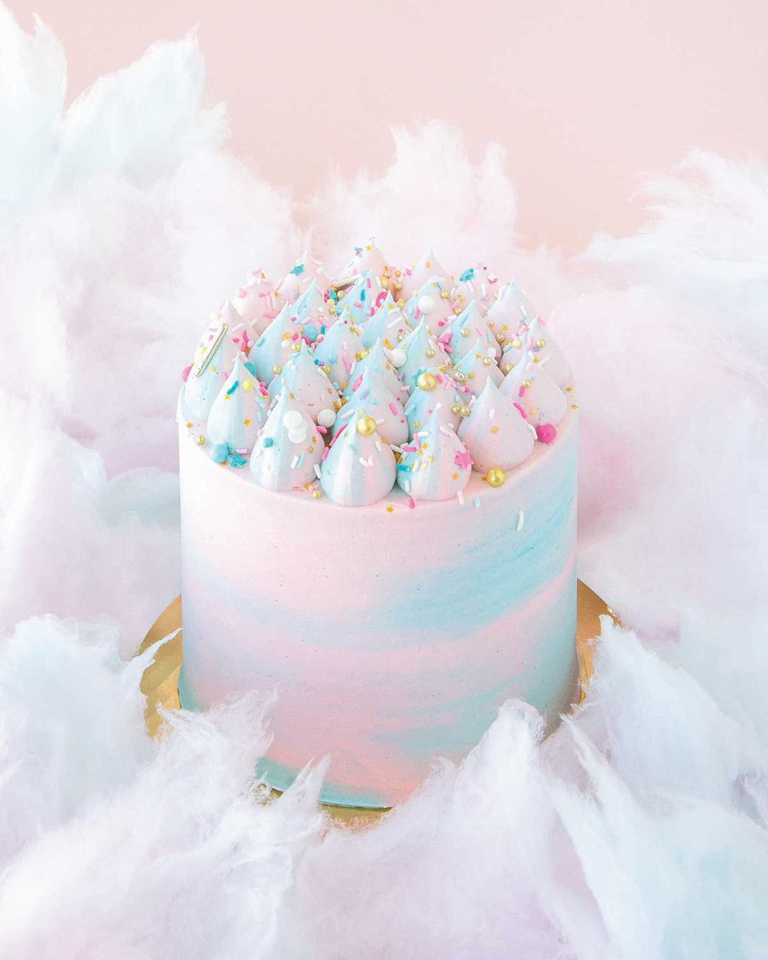 The Cotton Candy Cake by Jenna Rae Cakes features swirled pink and blue whipped frosting, colorful sprinkles, and fluffy cotton candy accents, set against a pale pink background.