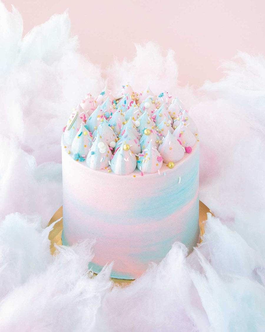 The Cotton Candy Cake by Jenna Rae Cakes features swirled pink and blue whipped frosting, colorful sprinkles, and fluffy cotton candy accents, set against a pale pink background.