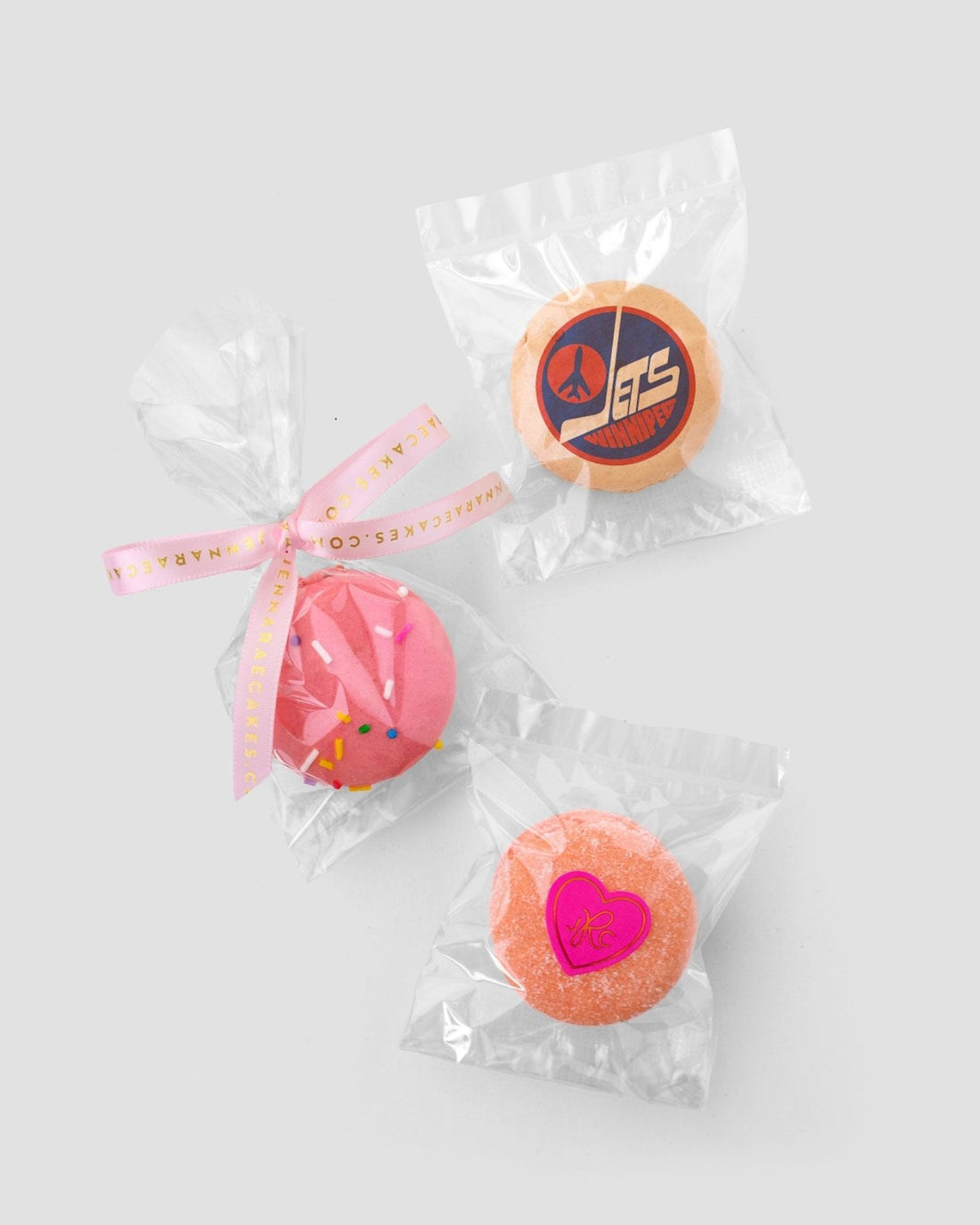 The Custom Single Macaron Bag by Jenna Rae Cakes includes three individually wrapped round macarons: one with a pink heart, one with pink icing and sprinkles, and one with an orange Jets logo on a blue background.